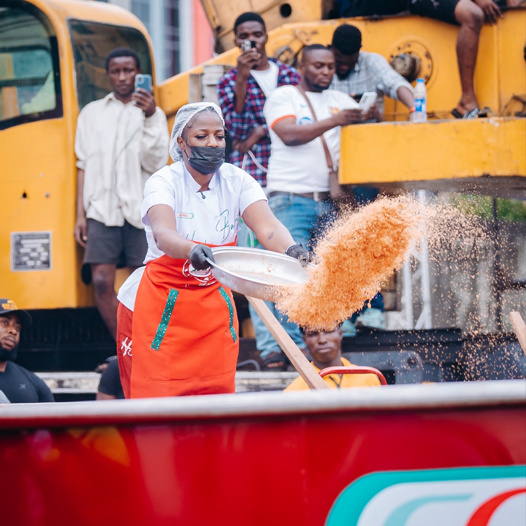 Hilda Baci wearing a face mask and an orange apron while tipping ingredients from a large metal pan into a massive cooking pot during her second successful record run. Photo Credit: Guinness World Records/Instagram